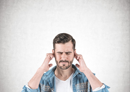 Portrait of bearded young man wearing jeans shirt and thinking hard with eyes closed and fingers on temples standing near concrete wall.の写真素材