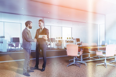 Businessmen in corner of office conference room with white and glass walls, brown carpet on the floor and long wooden table with beige chairs. White bookcase near the wall. Toned imageの写真素材