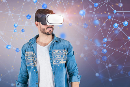 Portrait of bearded young man in jeans shirt wearing VR glasses standing over blurred blue purple...