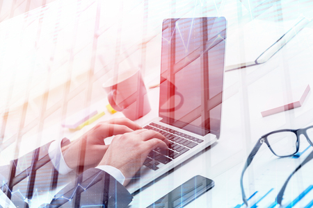 Hands of businessman in suit typing on laptop in office with double exposure of skyscraper and graph. Stock market concept. Toned imageの写真素材