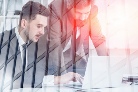 Two bearded young businessmen working with laptop in white office with double exposure of skyscraper. Concept of office work. Toned imageの写真素材