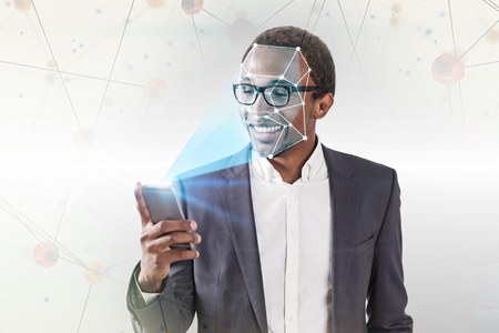 Smiling African American businessman in glasses using smartphone with face recognition technology over white background with polygons. Toned image double exposureの写真素材