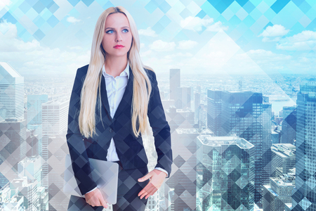 Pensive blonde businesswoman in black suit standing with laptop computer over cityscape background. Concept of woman in businessの写真素材