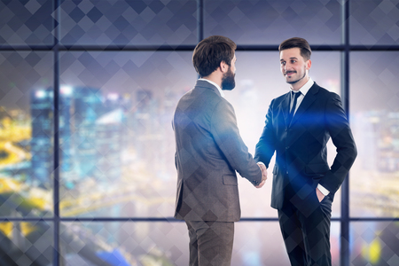 Two young businessmen in suits shaking hands standing in night panoramic office with blurred cityscape. Concept of partnership. Toned imageの写真素材
