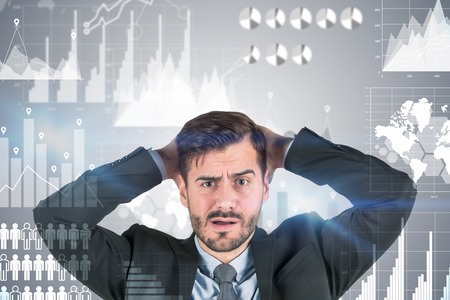 Confused young businessman with beard wearing dark suit and looking forward standing over gray...