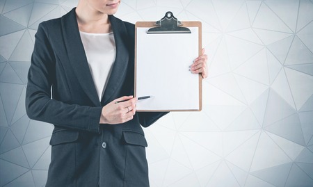 Portrait of unrecognizable young businesswoman wearing black suit and pointing at her clipboard with pen standing near geometric pattern wall. Mock upの写真素材