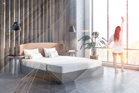 Woman standing in corner of master bedroom with concrete and wooden walls, concrete floor, double bed with two bedside tables, potted plant and stylish lamps. Toned image double exposureの写真素材