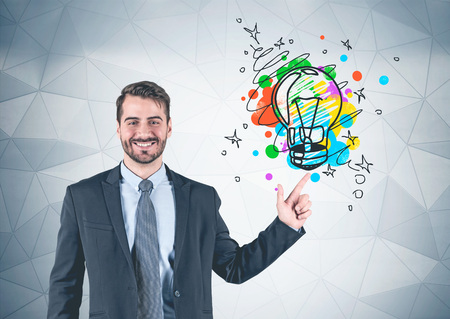 Portrait of smiling young bearded businessman wearing dark suit and pointing with his finger at geometric pattern wall with colorful light bulb sketch on it. Concept of bright ideaの写真素材