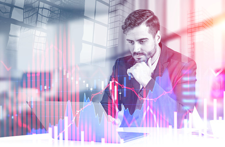 Serious young businessman working with laptop at office table over skyscraper background with double...