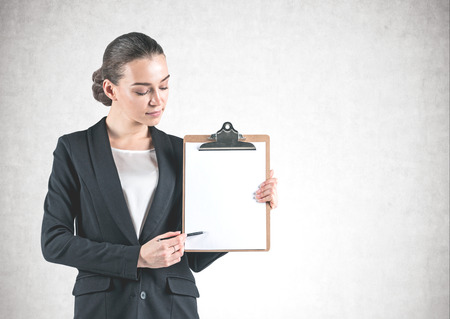 Portrait of young businesswoman wearing black suit and pointing at her clipboard with pen standing near concrete wall. Mock upの写真素材