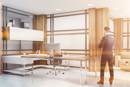 Businessman in corner of manager office with white striped walls, tiled floor, white and wooden computer table, white shelves and lounge area with sofa. Toned imageの写真素材