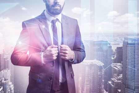Unrecognizable young businessman with beard wearing dark suit and standing over cityscape background with double exposure of network interface. Toned imageの写真素材
