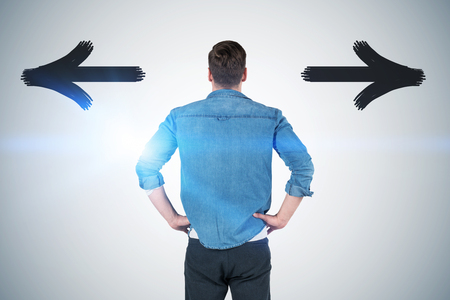 Rear view of caucasian young man in jeans shirt and gray pants looking at concrete wall with two arrows showing different directions. Concept of choice. Toned imageの写真素材