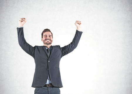 Portrait of smiling young businessman celebrating victory and standing with arms in the air near concrete wall. Mock upの写真素材