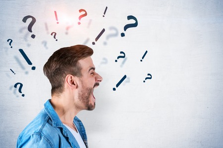 Side view of young man with brown hair wearing casual clothes and screaming. Concrete wall background with question and exclamation marks. Concept of communication problem. Mock upの写真素材