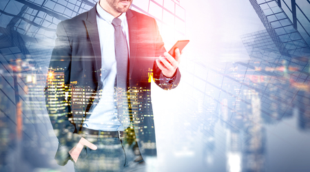 Unrecognizable businessman looking at smartphone screen with double exposure of night cityscape and skyscrapers. Communication concept. Toned imageの写真素材