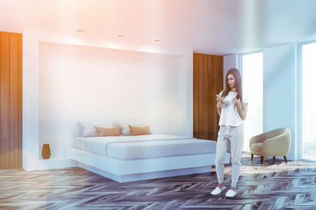 Young woman with smartphone standing in stylish bedroom interior with white walls, master bed and armchair. Toned imageの写真素材