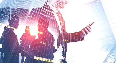 Portrait of business leader with smartphone with double exposure of his colleagues over skyscraper...