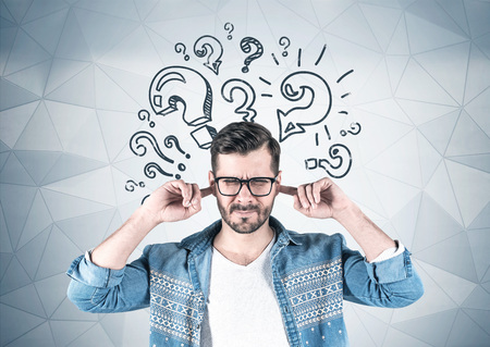 Young man in glasses and casual clothes covering his ears standing near gray wall with question marks drawn on it. Concept of decision makingの写真素材