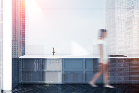 Young woman walking in minimalistic kitchen interior with white walls, dark wooden floor and gray countertop with built in sink and oven. Toned image double exposure blurredの写真素材