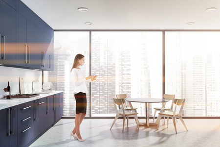 Woman with smartphone in suit standing in panoramic kitchen interior with round table and dark blue countertops. Toned imageの写真素材