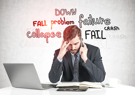 Concerned businessman talking on smartphone at office table near concrete wall with failure words written on it. Concept of crisis. Toned imageの写真素材