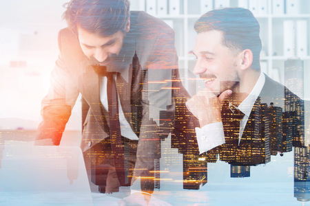 Two cheerful handsome businessmen working with laptop in modern office with double exposure of night cityscape. Concept of management. Toned imageの写真素材