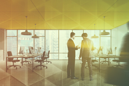 Two young businessmen discussing documents in modern panoramic open space office with yellow ceiling, white walls and rows of computer tables. Toned image double exposure blurの写真素材