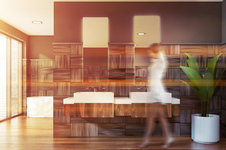 Young woman walking in stylish bathroom with gray and wooden walls, wooden floor, double sink and white bathtub. Toned image blurの写真素材