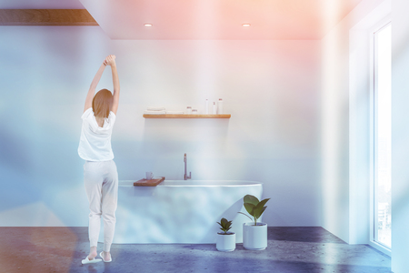 Rear view of woman in pajamas standing in comfortable bathroom with white walls and white bathtub. Toned imageの写真素材