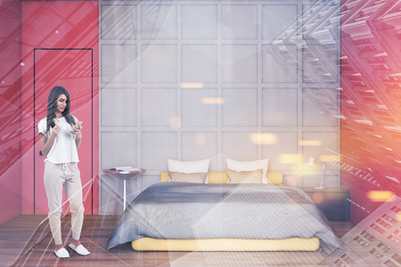 Young woman with smartphone standing in modern bedroom interior with white and red walls, wooden floor and gray master bed. Toned image double exposureの写真素材