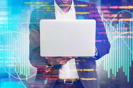 Unrecognizable young African American man in suit looking at laptop. Blurred background with lines of code and graphs. Concept of hi tech startup and software development. Toned image double exposureの写真素材