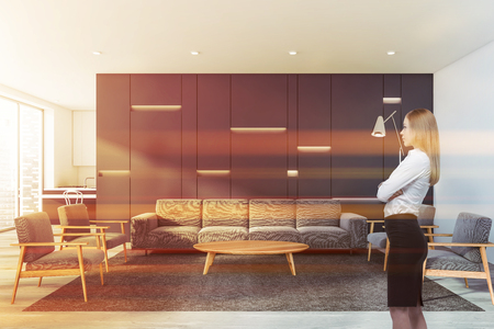 Blonde young woman standing in modern living room with gray and white walls, sofa and armchairs near coffee table and kitchen in background. Concept of real estate agent work. Toned imageの写真素材