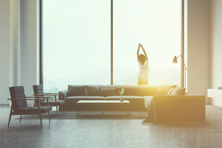 Young woman in casual clothes standing in luxury panoramic living room with long gray sofa, comfortable armchairs and wooden coffee table. Toned image double exposureの写真素材