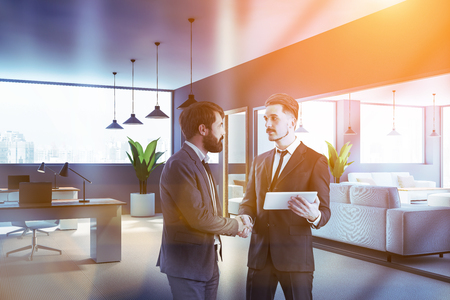 Two young businessmen shaking hands in modern office with open space area and waiting room with couches. Concept of partnership. Toned imageの写真素材