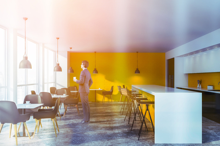 Young businessman in glasses standing in modern coffee shop interior with bright yellow walls, bar counter and square white tables with black chairs. Toned imageの写真素材
