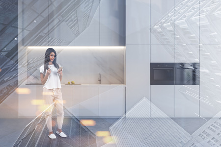 Young woman with coffee and smartphone standing in modern kitchen interior with marble walls, white countertops and cupboards and two ovens. Toned image double exposureの写真素材