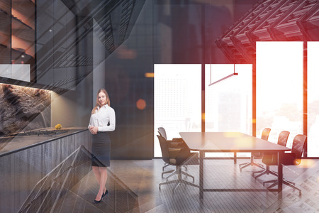 Young woman with blond hair standing in modern kitchen interior with gray cupboards and countertops and long dining table with chairs. Toned image double exposureの写真素材