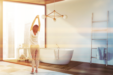 Woman in pajamas standing in luxury bathroom with panoramic windows, white walls, comfortable bathtub and ladder with towel on it. Toned imageの写真素材