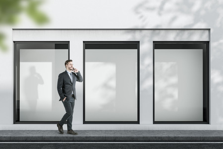 Smiling businessman with phone walking near white building with three vertical mock up posters in windows. Concept of advertising and marketing.の写真素材