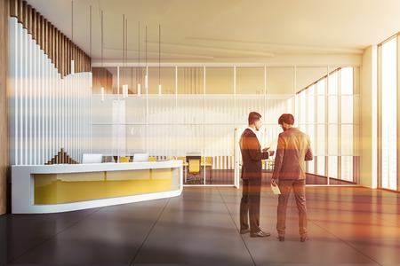 Two businessmen discussing documents in modern office hall with yellow reception desk and open space area in background. Toned imageの写真素材