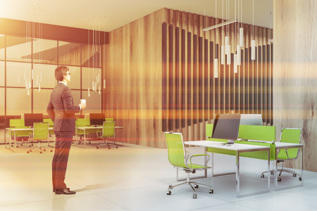 Handsome young businessman in glasses standing in modern open space office with gray and wooden walls and rows of white and green computer tables. Toned imageの写真素材