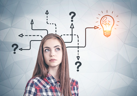 Thoughtful young woman in casual clothes standing near geometric pattern wall with question marks and lightbulb drawn on it. Concept of good ideaの写真素材