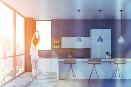 Rear view of young woman standing in luxury panoramic kitchen interior with white marble walls, gray countertops and wooden bar with stools. Toned imageの写真素材
