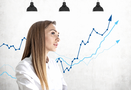 Thoughtful blonde businesswoman in white shirt standing near concrete wall with growing graph drawn on it. Concept of financial success and stock marketの写真素材