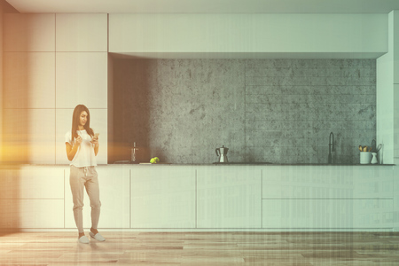 Young woman with smartphone and coffee standing in minimalistic kitchen with concrete walls and white countertops with built in sink and cooker. Toned image double exposureの写真素材
