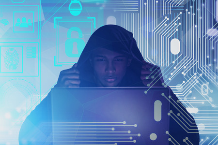 Serious African American hacker in hoodie looking at laptop. Double exposure of circuit and online...
