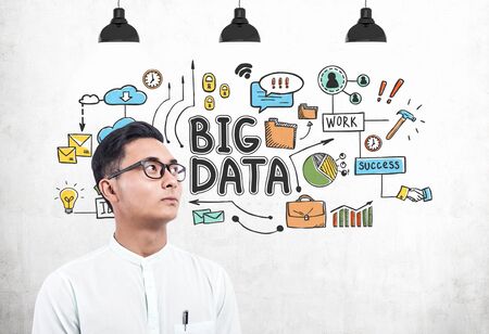 Portrait of serious young Asian businessman in glasses and white shirt standing near concrete wall with colorful big data sketch drawn on it.の写真素材