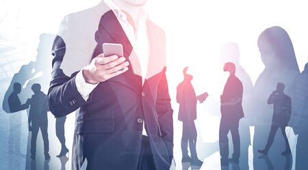 Unrecognizable businessman with phone and double exposure of his colleagues over abstract skyscraper background. Concept of teamwork. Toned imageの写真素材