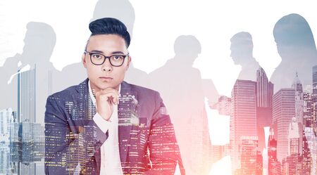 Thoughtful Asian businessman in glasses looking at camera. Double exposure of cityscape and silhouettes of business people. Concept of leadership. Toned imageの写真素材